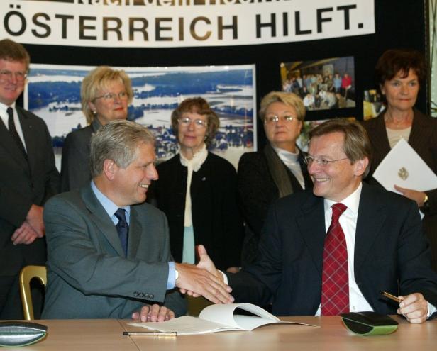 Zwei Männer im Anzug geben sich die Hand vor einer Gruppe von Personen und einem Banner „Österreich hilft“.