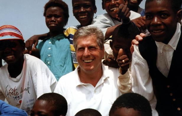 Ein lächelnder Mann mit grauen Haaren posiert mit einer Gruppe afrikanischer Kinder.