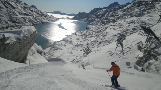 Skifahrer auf einer schneebedeckten Piste mit einem Bergsee im Hintergrund.