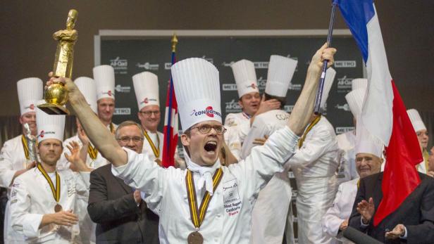 Thibaut Ruggeri feiert seinen Sieg beim Bocuse d'Or mit einer Trophäe und der französischen Flagge.