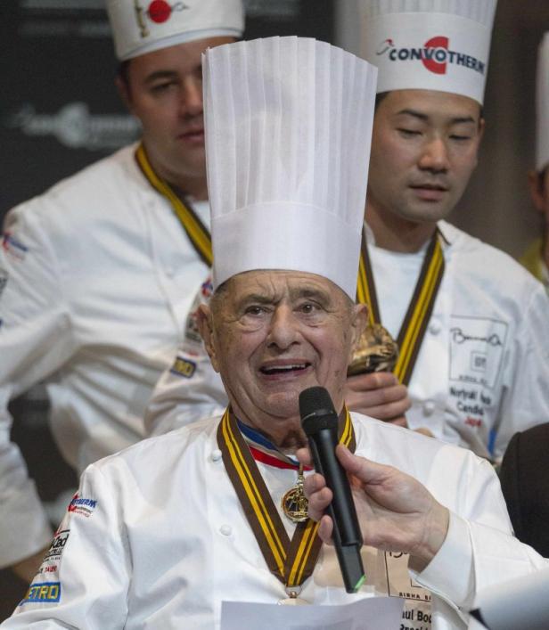 Paul Bocuse, ein Koch mit Kochmütze und Medaillen, spricht in ein Mikrofon.