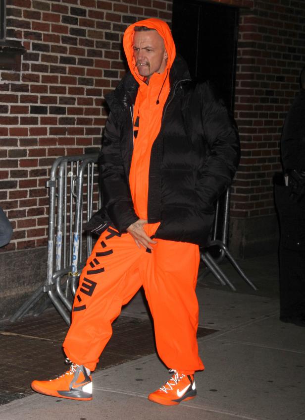 Ein Mann in einem orangefarbenen Trainingsanzug und einer schwarzen Jacke steht draußen.