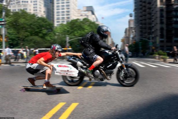 Ein Skateboardfahrer hält sich an einem Motorrad fest und transportiert Pizzakartons.