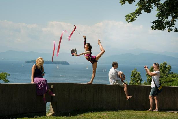 Eine Turnerin balanciert mit einem Buch und einem Band auf einer Mauer vor einer Seenlandschaft.