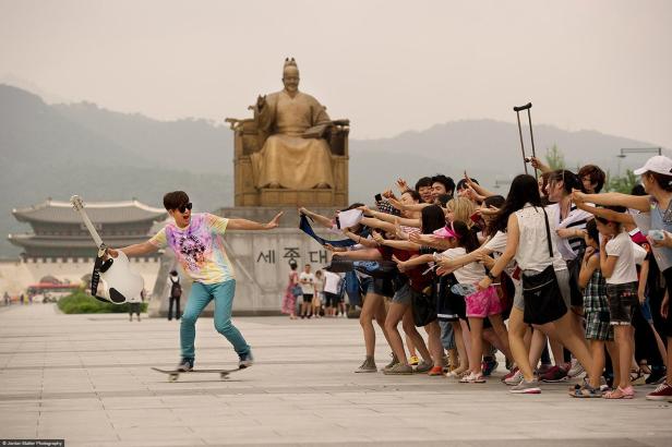 Ein Mann fährt mit einem Skateboard und einer Gitarre vor einer Statue, während Fans ihre Hände ausstrecken.