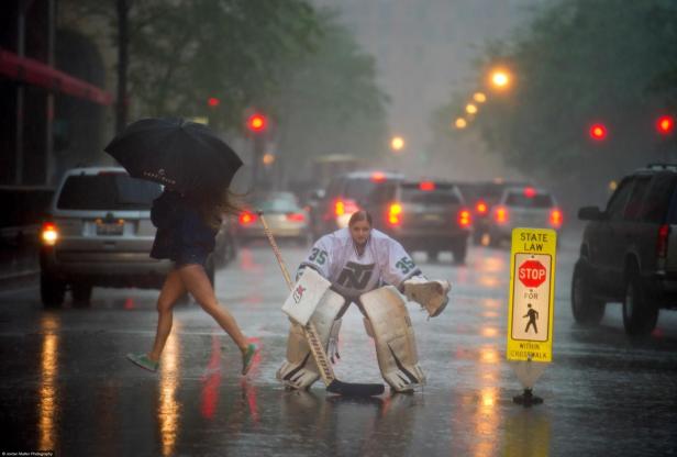 Eine Frau in Hockeyausrüstung steht auf einer regnerischen Straße, während eine andere Frau mit einem Regenschirm vorbeigeht.