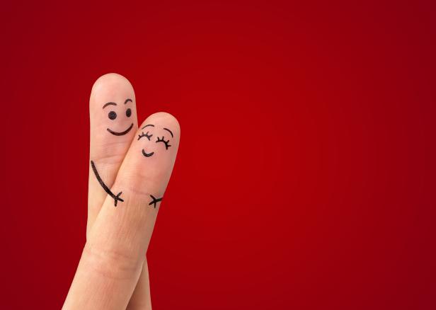 Zwei Finger mit aufgemalten Gesichtern umarmen sich vor rotem Hintergrund.