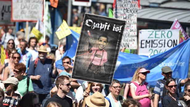 Demonstranten halten Schilder, darunter eines mit einem Bild von Barack Obama in Tracht und der Aufschrift „O’zapft is!“.