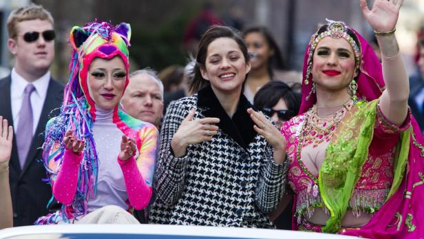 Marion Cotillard mit zwei extravagant gekleideten Personen auf einem Wagen.