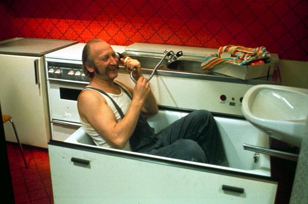 Ein Mann sitzt bekleidet in einer Badewanne und hält einen Duschkopf ans Ohr.