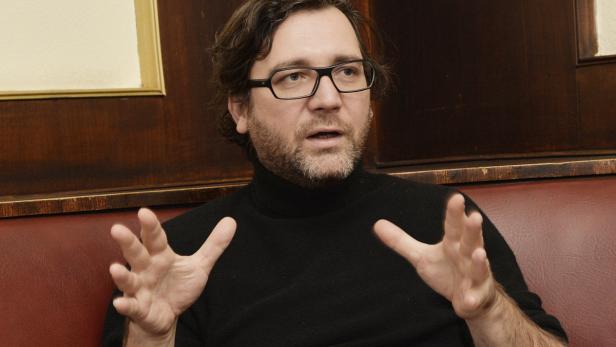 Ein Mann mit Brille und Bart gestikuliert vor einer dunklen Holzvertäfelung.