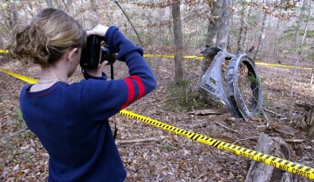 Eine Frau fotografiert in einem Wald ein Trümmerteil, das mit einem gelben Absperrband umgeben ist.