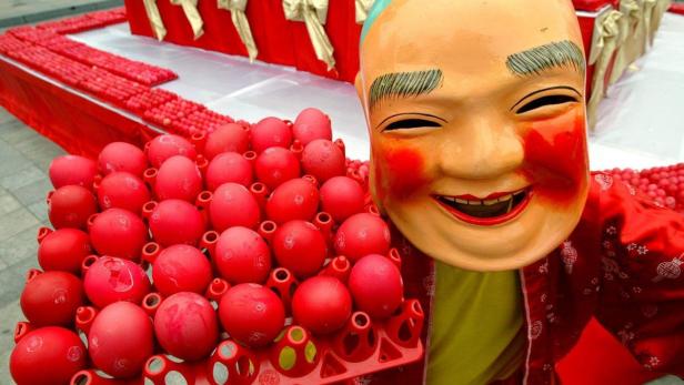 Eine Person mit einer traditionellen chinesischen Maske hält eine Eierpalette mit rot gefärbten Eiern.