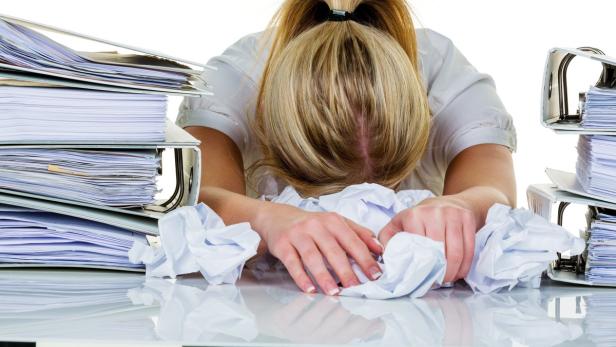 Eine erschöpfte Frau liegt mit dem Kopf auf einem Schreibtisch voller Papier und Ordner.