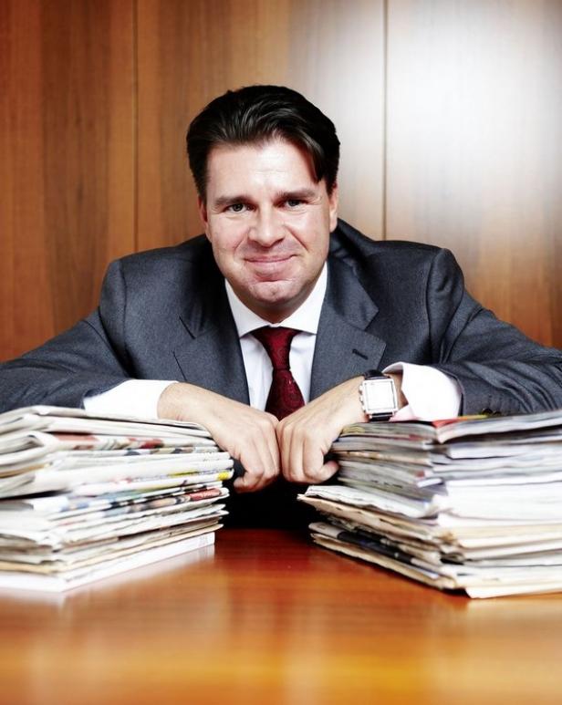 Ein Mann im Anzug sitzt an einem Tisch mit zwei Stapeln Zeitungen.