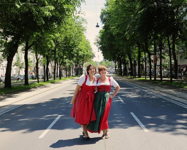 Zwei Frauen in Dirndln stehen auf einer von Bäumen gesäumten Straße.