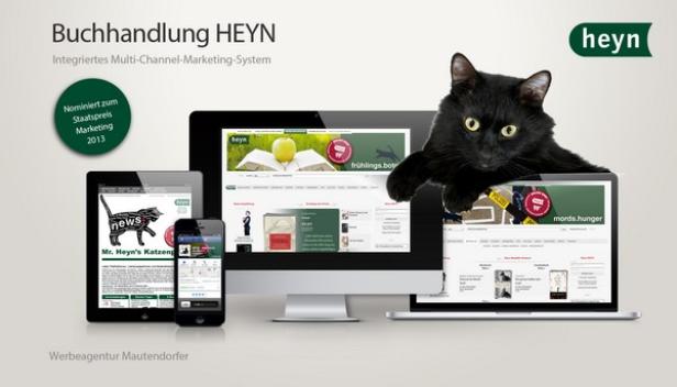 Eine schwarze Katze schaut über einen Laptop-Bildschirm, der die Webseite der Buchhandlung Heyn zeigt.