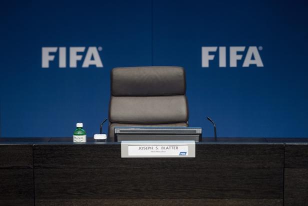 Ein leerer Stuhl mit dem Namensschild „Joseph S. Blatter, FIFA Präsident“ vor einem FIFA-Hintergrund.