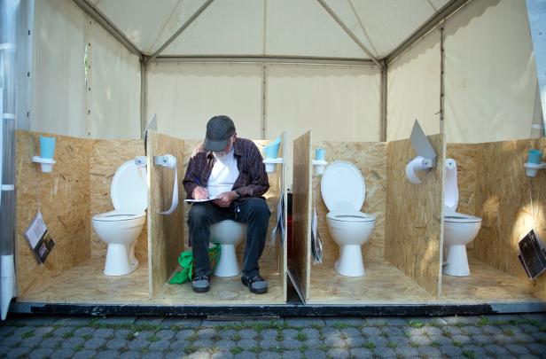 Ein Mann sitzt auf einer Toilette in einer Kabine und schreibt etwas auf ein Blatt Papier.