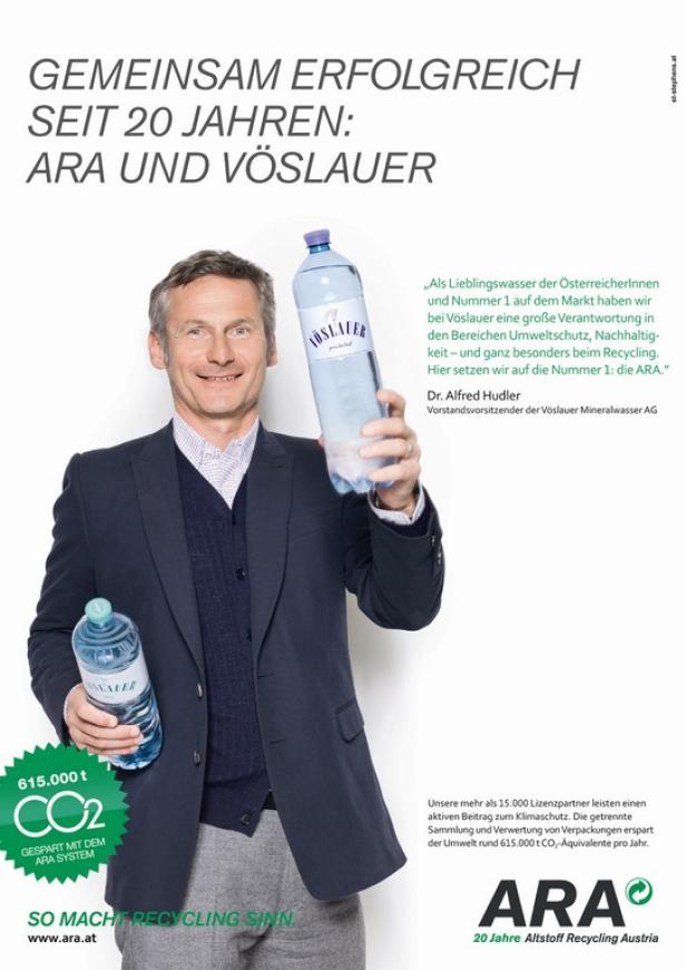 Ein Mann im Anzug präsentiert zwei Flaschen Vöslauer Mineralwasser vor einem Werbeplakat für ARA und Vöslauer.