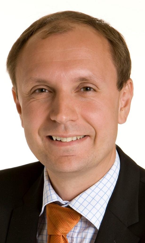 Ein lächelnder Mann mit Anzug und orangefarbener Krawatte vor einem weißen Hintergrund.
