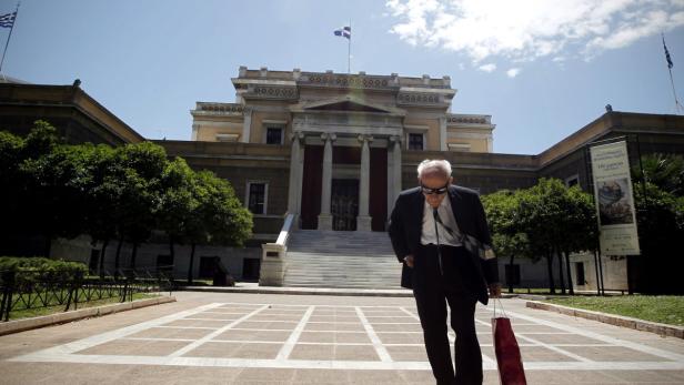 Ein Mann mit Anzug und Sonnenbrille geht vor einem Gebäude mit griechischer Flagge.