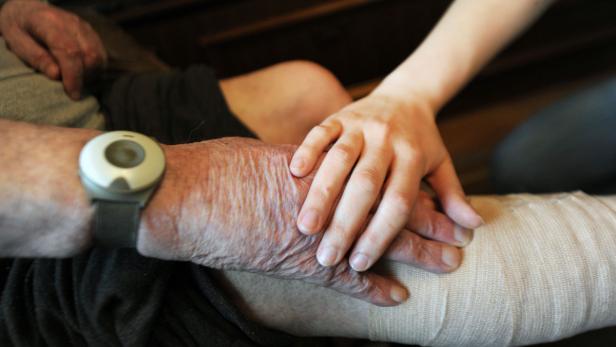 Eine junge Hand liegt auf der Hand einer älteren Person mit einem medizinischen Armband.