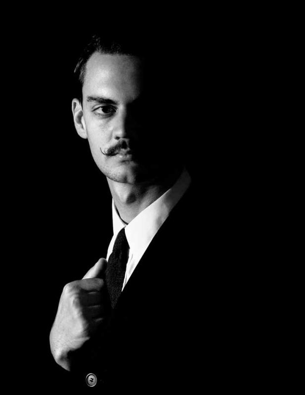 Ein Mann mit Schnurrbart im Anzug, zur Hälfte im Schatten.