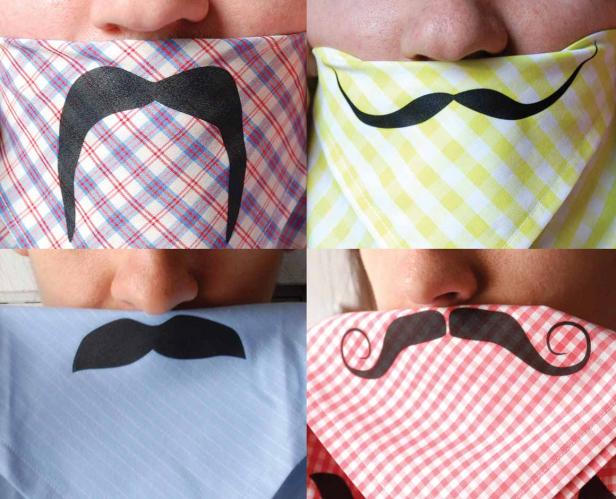 Vier Personen tragen Tücher mit aufgedruckten Schnurrbärten vor dem Gesicht.