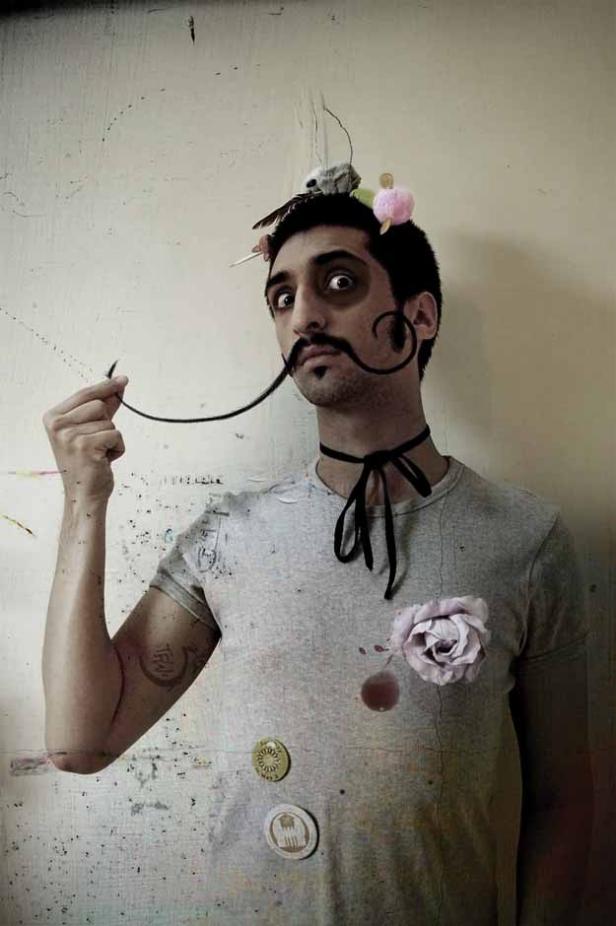 Ein Mann mit aufgemaltem Schnurrbart und ungewöhnlicher Kopfbedeckung blickt in die Kamera.