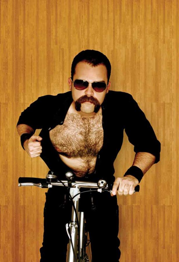 Ein Mann mit Sonnenbrille, Schnurrbart und geöffnetem Hemd fährt Fahrrad.