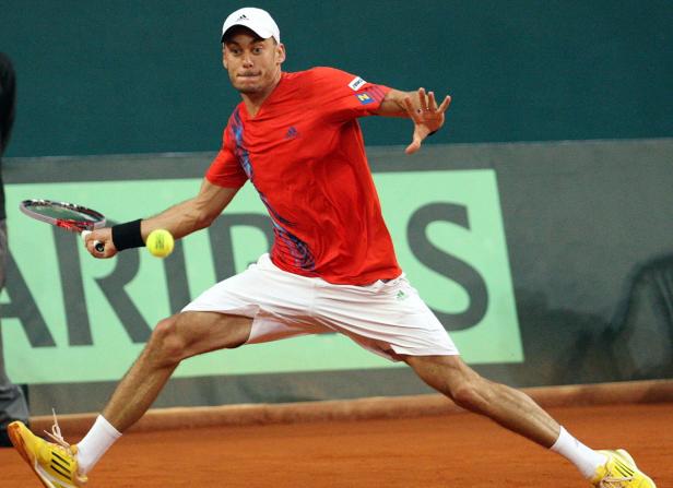 Ein Tennisspieler in rotem Hemd und weißer Hose schlägt einen Ball.