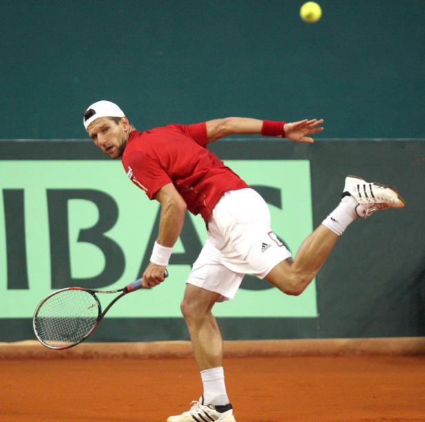 Ein Tennisspieler in rotem Hemd und weißer Hose schlägt einen Ball.