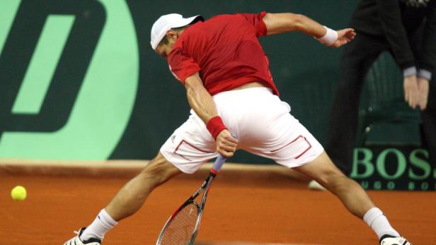 Ein Tennisspieler in rotem Hemd und weißer Hose schlägt einen Ball.