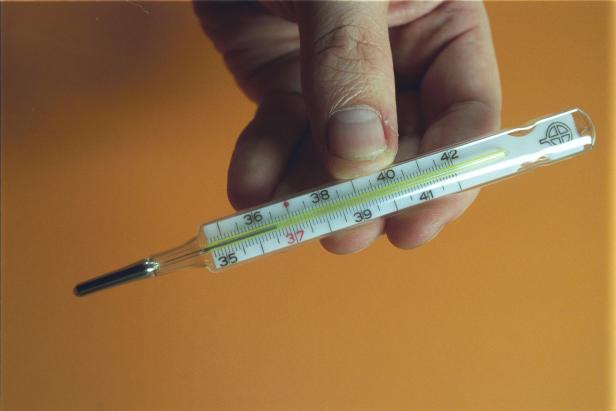 Eine Hand hält ein Quecksilberthermometer, das eine Temperatur von etwa 37,5 Grad Celsius anzeigt.
