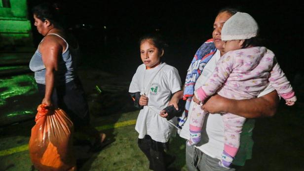 Eine Frau trägt ein Baby, während ein Mädchen und eine andere Frau mit einer Tüte im Dunkeln gehen.