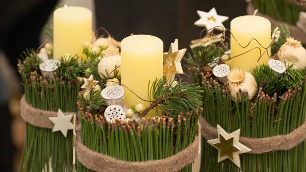 Oh du blühende Adventzeit Drei Adventsgestecke mit gelben Kerzen, Tannenzweigen und Sternen verziert.