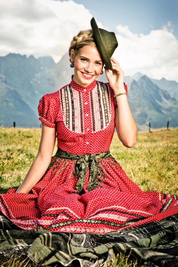 Eine Frau in Tracht sitzt auf einer Decke vor einer Bergkulisse.