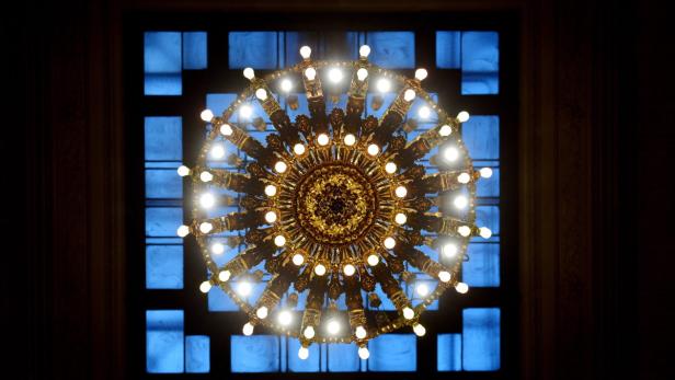 Ein detailreicher, goldener Kronleuchter hängt vor einem Fenster mit bläulichen Scheiben.