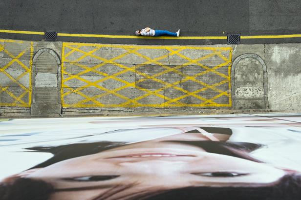 Eine Person liegt auf dem Bürgersteig vor einem großen Porträt einer Frau.