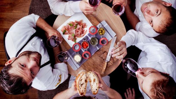 Eine Gruppe von Personen sitzt an einem Tisch mit Wein, Brot, Aufschnitt und Dips.