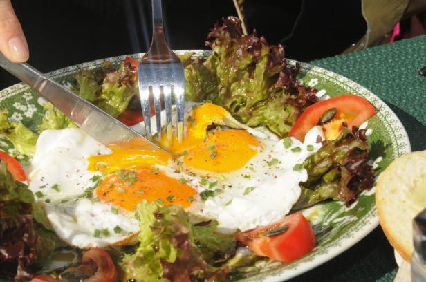Ein Teller mit Spiegeleiern, Salat und Tomaten wird mit Messer und Gabel gegessen.