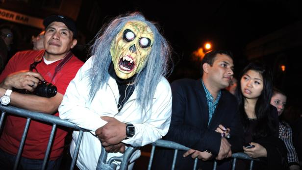 Eine Person mit einer gruseligen Zombie-Maske lehnt an einer Absperrung, umgeben von anderen Menschen.