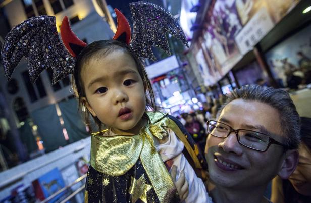 Ein kleines Mädchen mit Teufelshörnern und Umhang wird von einem Mann gehalten.