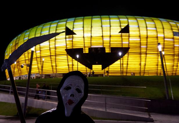 Eine Person in Halloween-Kostüm steht vor einem Gebäude, das wie ein Kürbis beleuchtet ist.