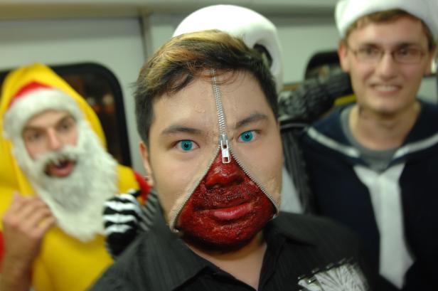 Ein Mann mit einem Reißverschluss-Make-up-Effekt im Gesicht, umgeben von Personen in Kostümen.