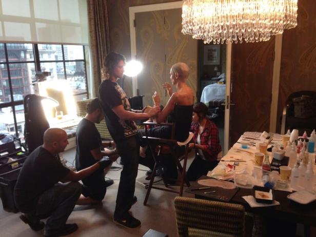 Visagisten arbeiten an einer Frau mit Glatze in einem Raum mit Kronleuchter.