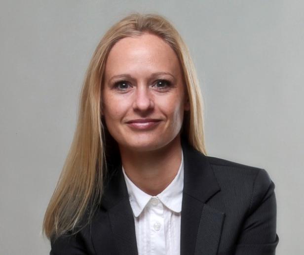 Eine blonde Frau in einem schwarzen Blazer und einer weißen Bluse lächelt.