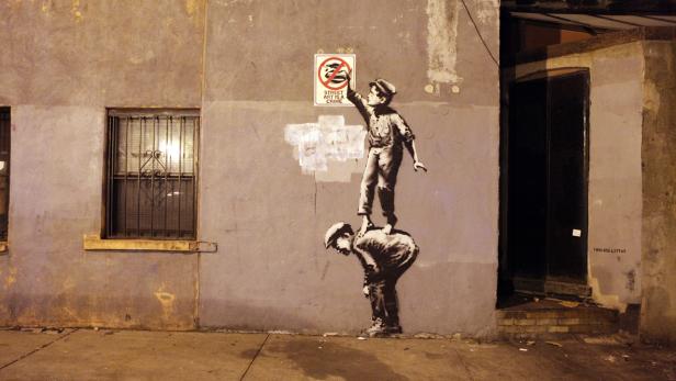 Ein Banksy-Graffiti zeigt einen Jungen, der auf den Schultern eines Mannes steht und ein „Street Art is a Crime“-Schild bemalt.