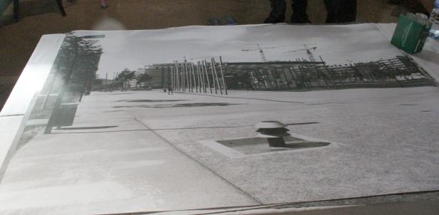 Eine Schwarzweißaufnahme eines Platzes mit modernen architektonischen Elementen und Baukränen im Hintergrund.